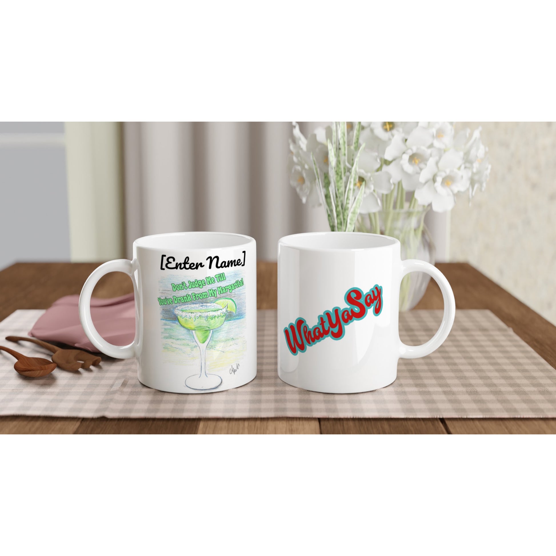 Two personalized white ceramic 11oz mugs with personalized original motto [Enter Name] Don’t Judge Me Till You’ve Drank From My Margarita on front and WhatYa Say logo on back dishwasher and microwave safe from WhatYa Say Apparel sitting on coffee table with green and white placemat. 