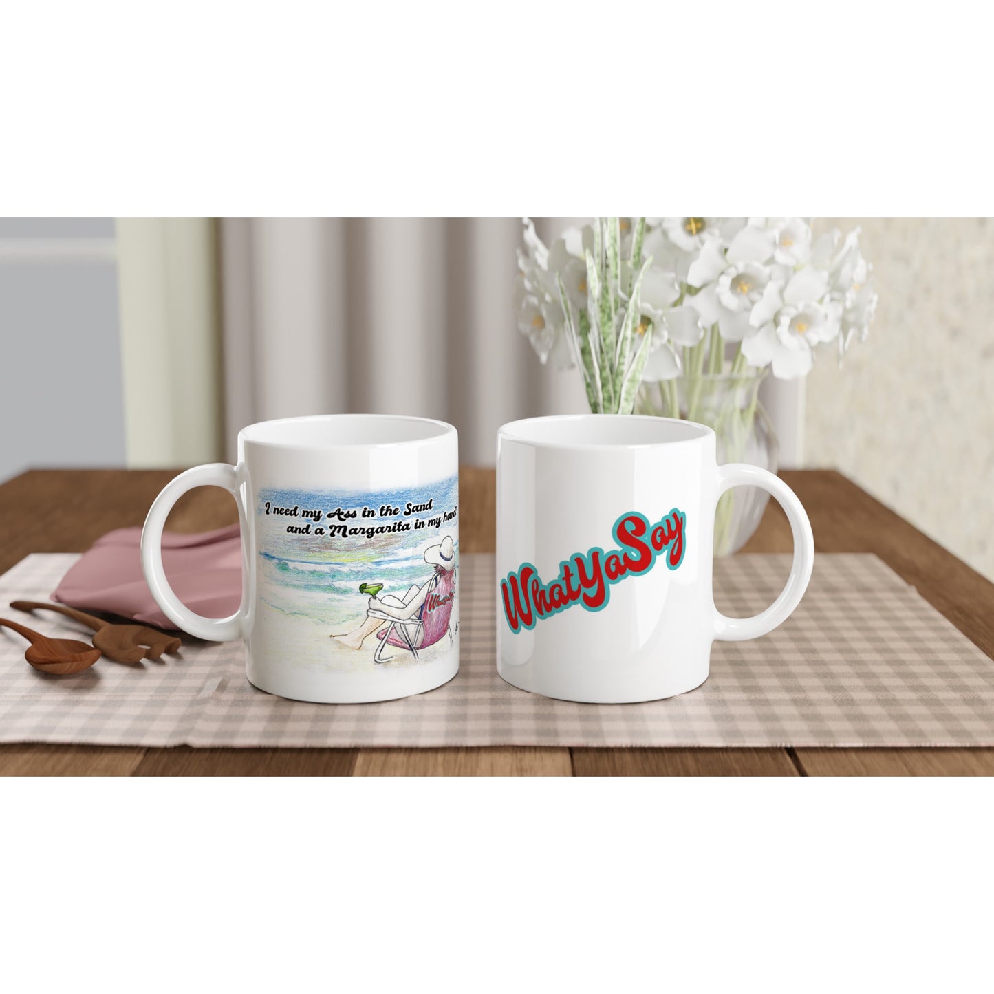 Two White ceramic 11oz mugs with motto I need my Ass in the Sand and a Margarita in my hand on front and WhatYa Say logo on back coffee mugs are dishwasher and microwave safe from WhatYa Say Apparel sitting on coffee table with green and white placemat.