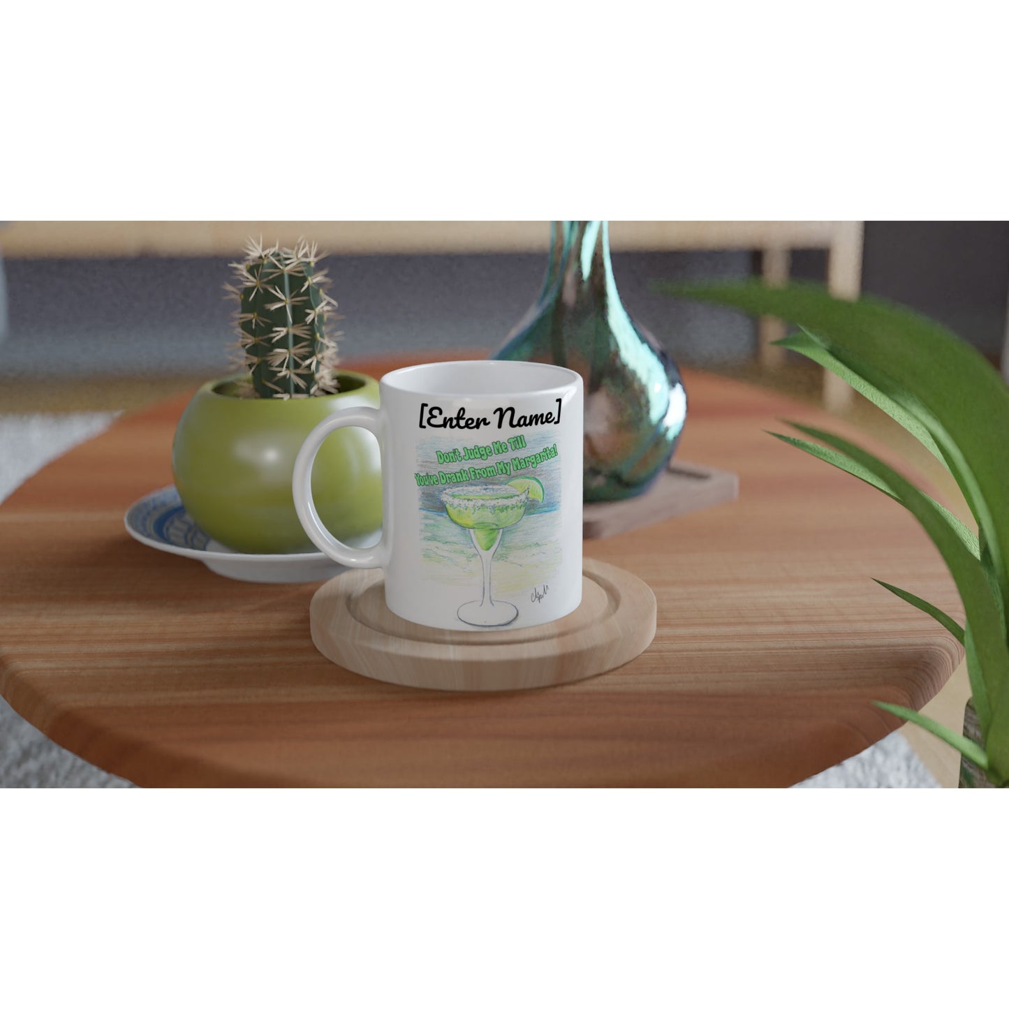 A personalized white ceramic 11oz mug with personalized original motto [Enter Name] Don’t Judge Me Till You’ve Drank From My Margarita on front and WhatYa Say logo on back dishwasher and microwave safe from WhatYa Say Apparel sitting on coaster on coffee table with green potted cactus and silver vase.
