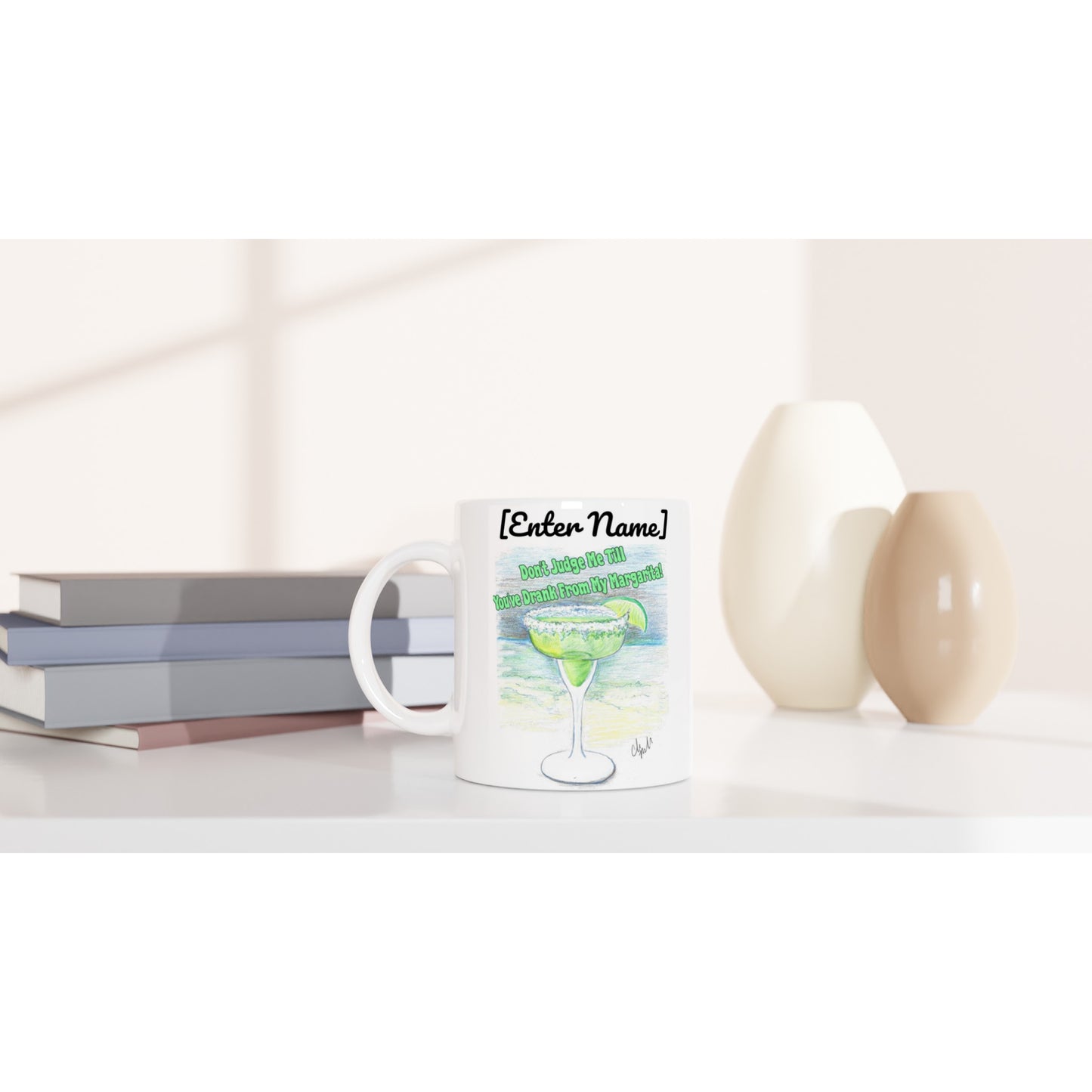 A personalized white ceramic 11oz mug with personalized original motto [Your Name] Don’t Judge Me Till You’ve Drank From My Margarita on front and WhatYa Say logo on back dishwasher and microwave safe from WhatYa Say Apparel sitting on coffee table with books and two vases.