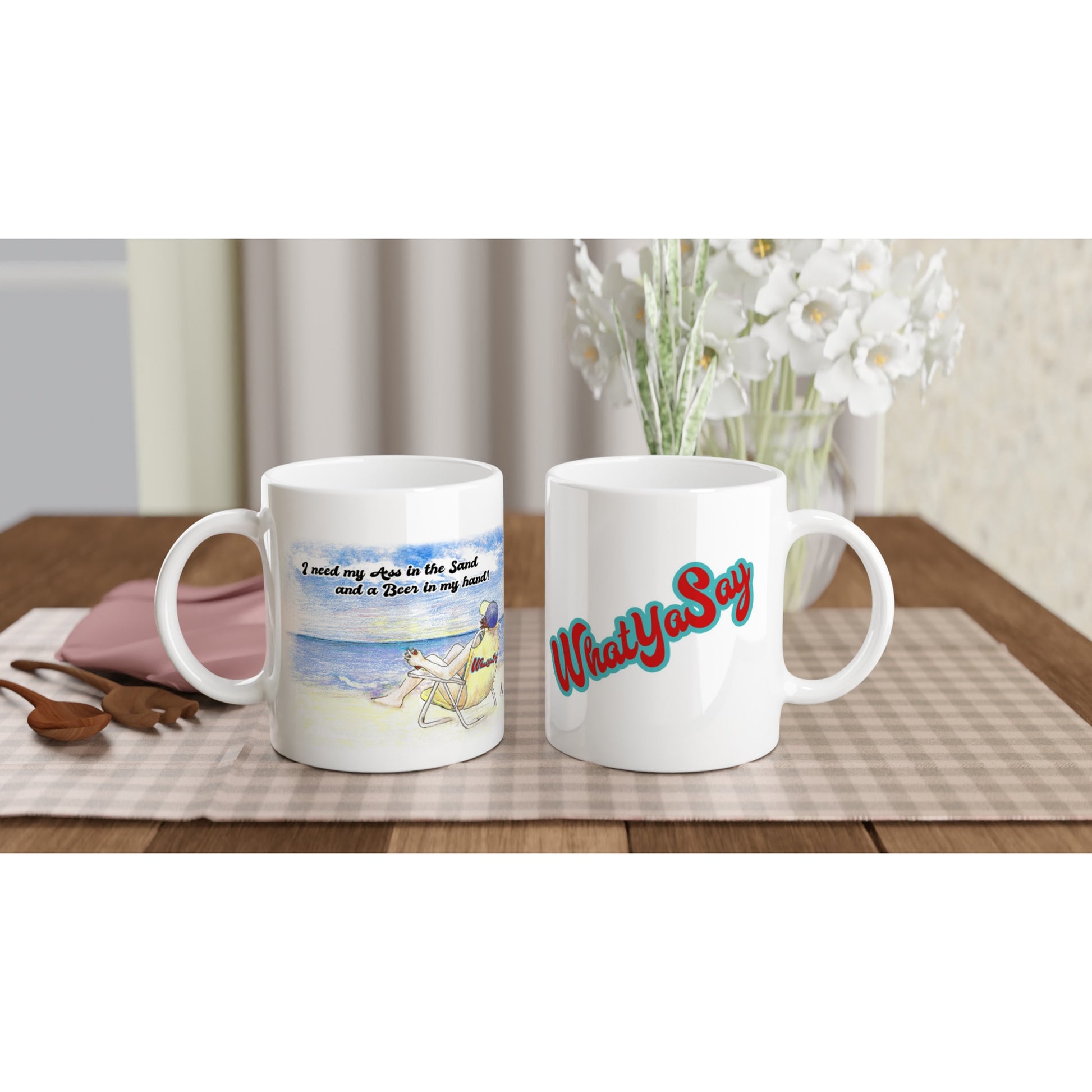 Two White ceramic 11oz mugs with motto I need my Ass in the Sand and a Beer in my hand on front and WhatYa Say logo on back coffee mugs are dishwasher and microwave safe from WhatYa Say Apparel sitting on coffee table with green and white placemat.