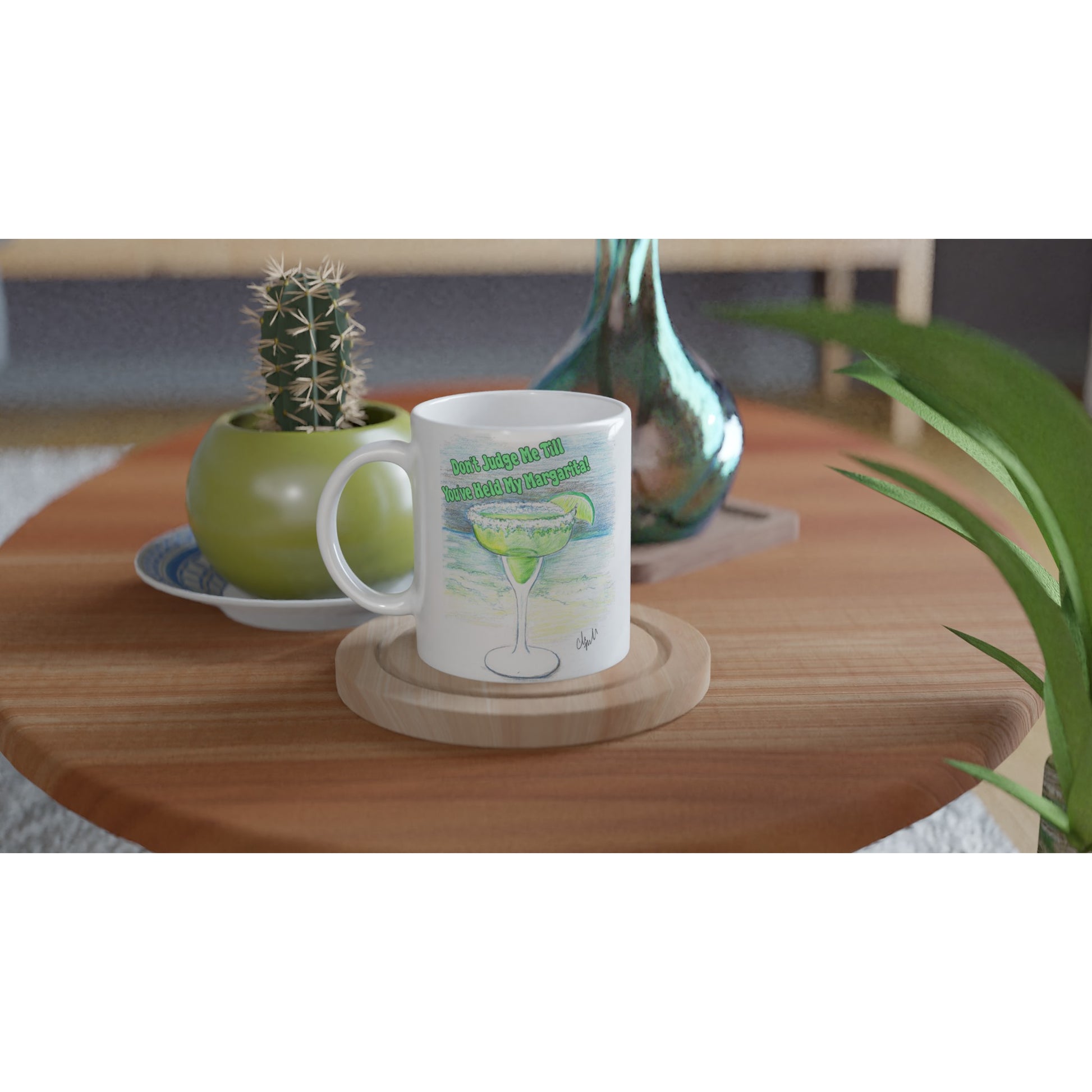 White ceramic 11oz mug with motto Don't Judge Me till You've Held my Margarita coffee mug dishwasher and microwave safe from WhatYa Say Apparel sitting on coaster on coffee table with green potted cactus and silver vase.