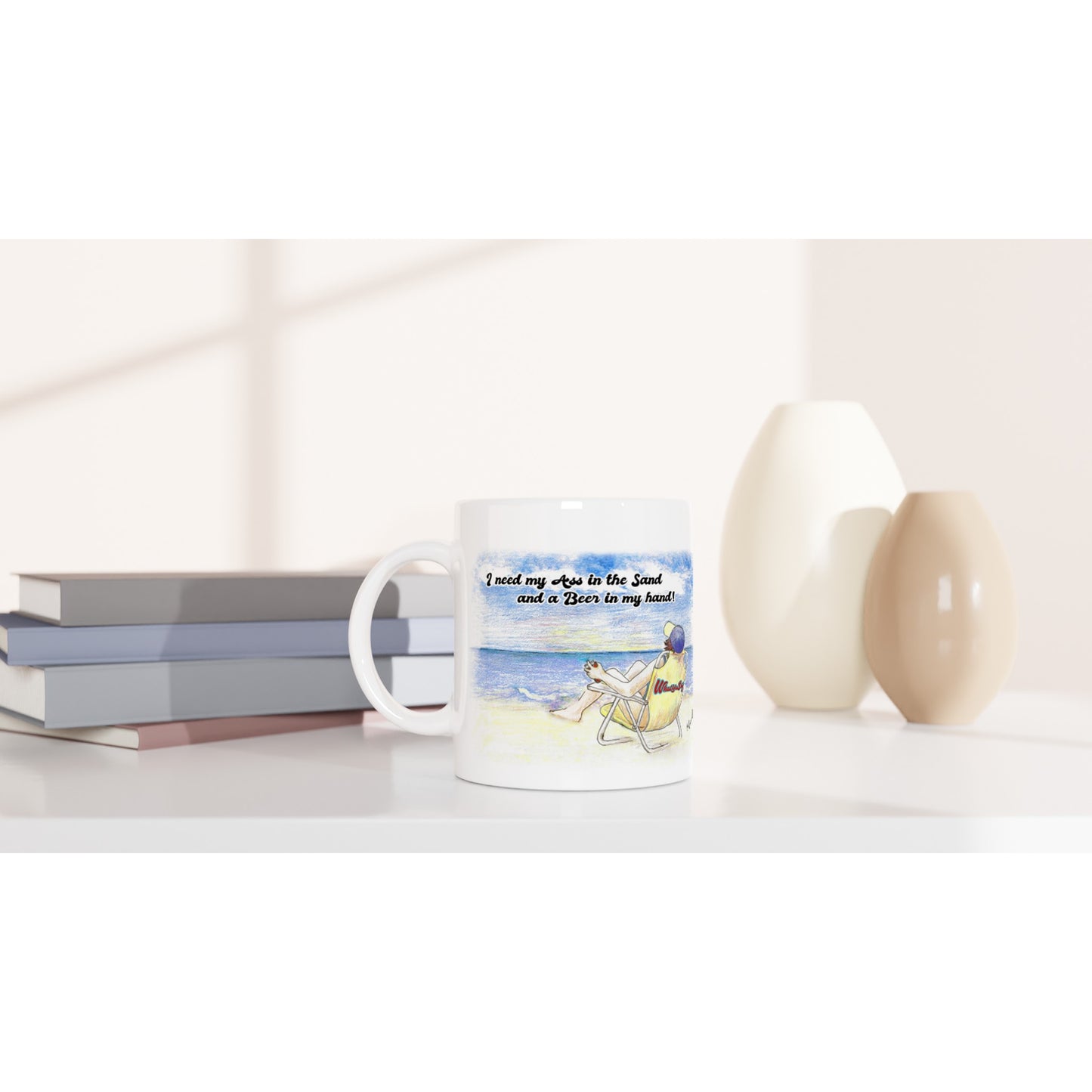 A white ceramic 11oz mug with motto I need my Ass in the Sand and a Beer in my hand on front and WhatYa Say logo on back dishwasher and microwave safe from WhatYa Say Apparel sitting on coffee table with books and two vases.