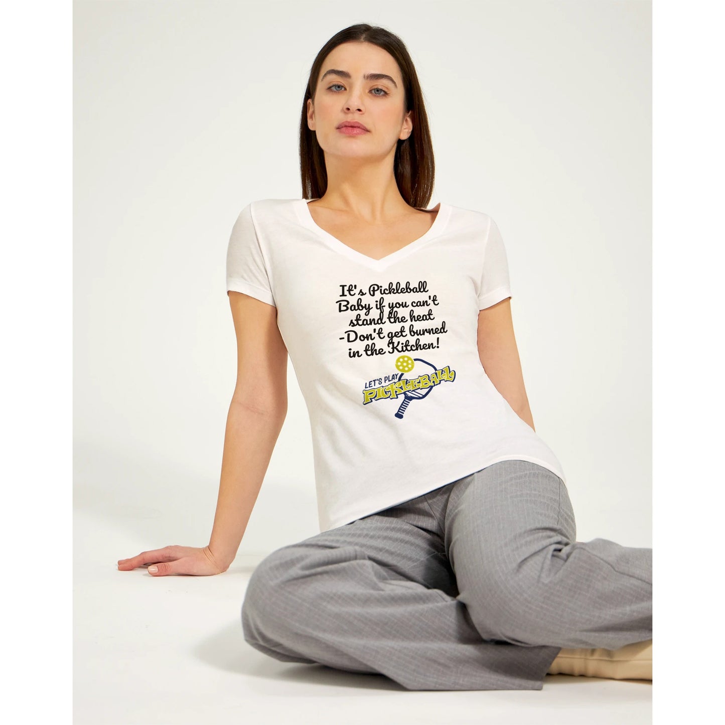 A premium women’s V-neck t-shirt from combed and ring-spun cotton with original logo It's PIckleball Baby if you can't stand the heat - Don't get burned in the Kitchen and Let's Play PickleBall on front from WhatYa Say Apparel worn by dark-haired Female model sitting on floor looking forward.