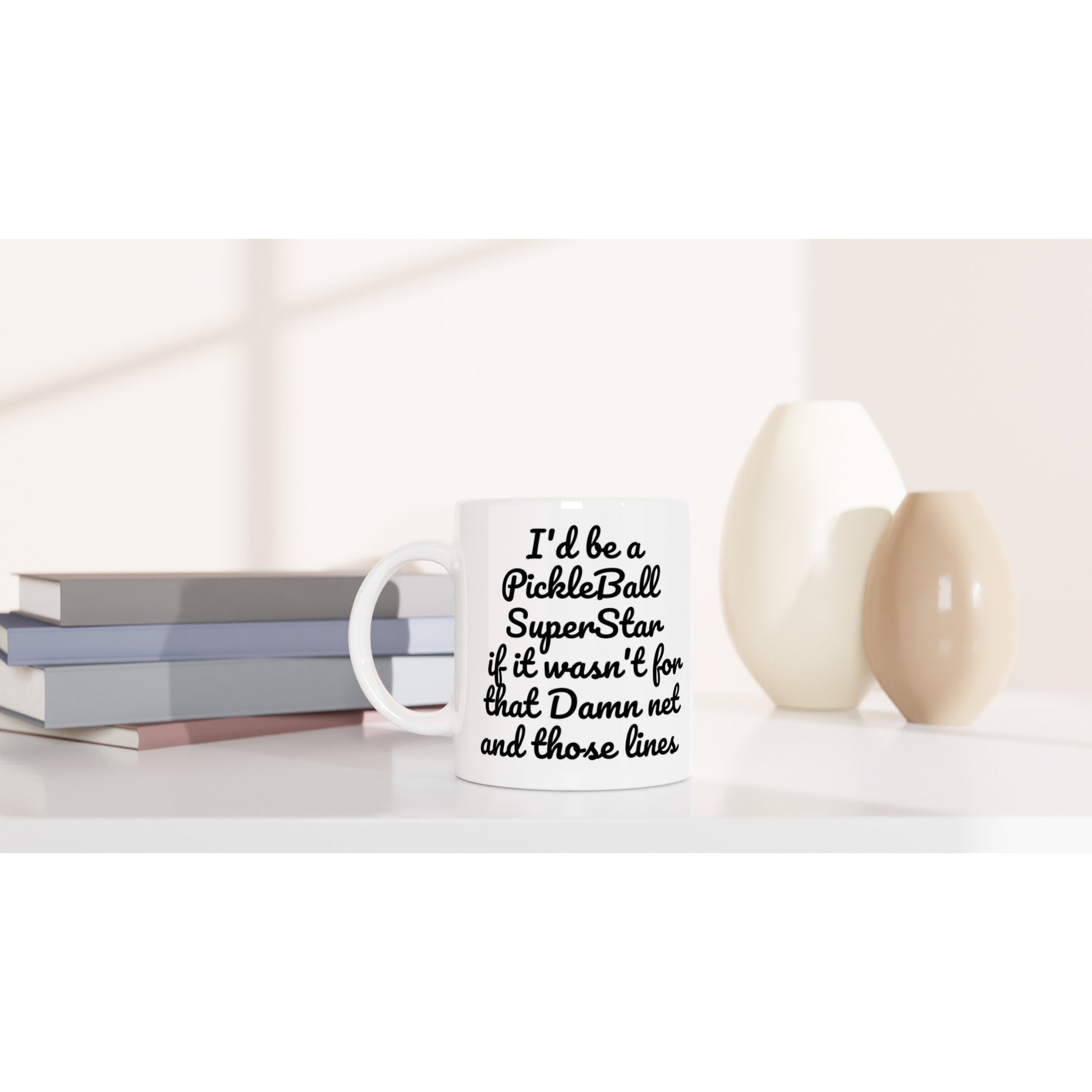 I'd be a PickleBall SuperStar if it wasn't for that Damn net and those lines Pickleball 11oz white ceramic mug and coffee mug is dishwasher safe and microwave safe sitting on table with books and vases.