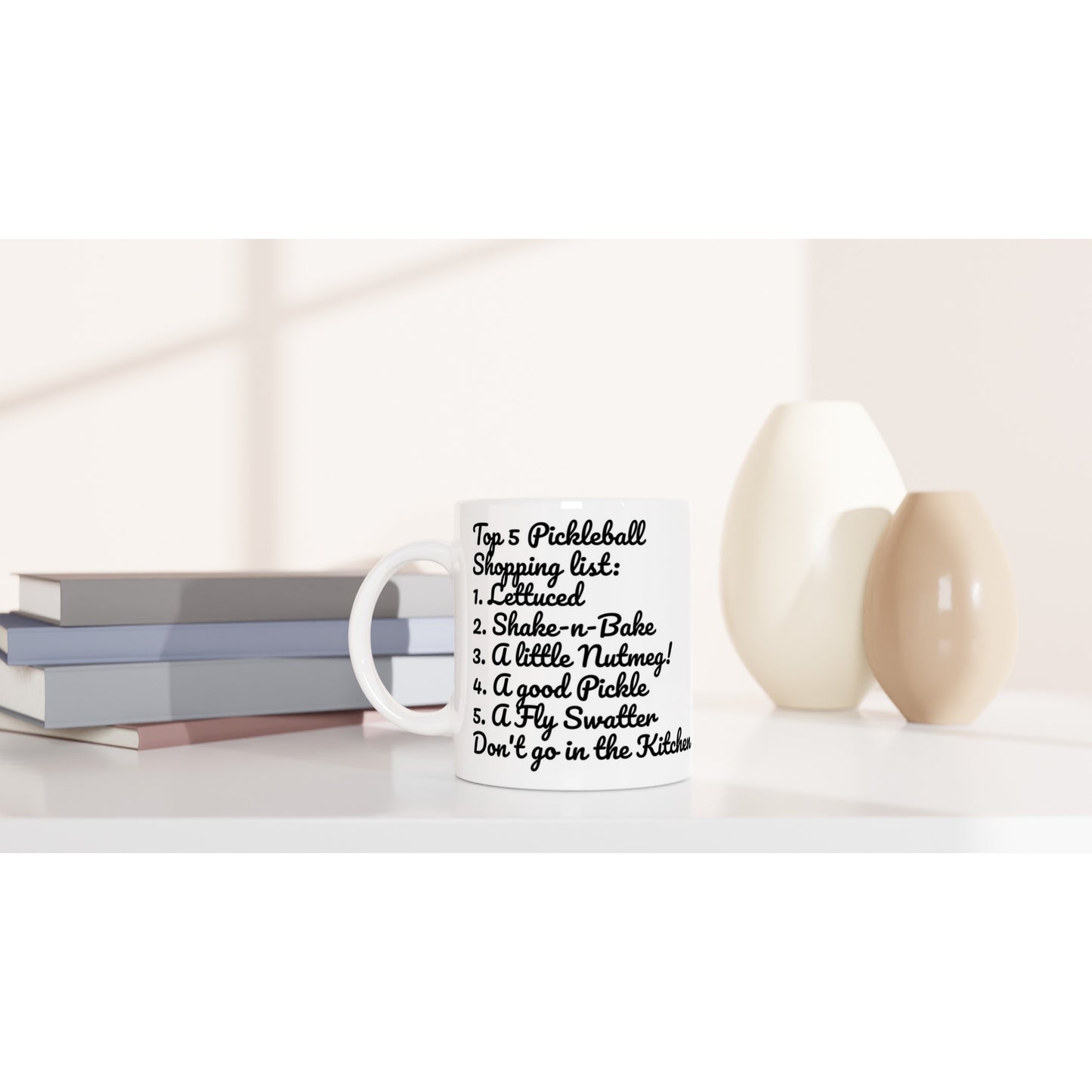 White ceramic 11oz mug with original motto Top 5 Pickleball Shopping list lettuce, Shake-n-bake, Nutmeg, Pickle, a Fly Swatter Don’t go in the kitchen on front and Let's Play Pickleball logo on back dishwasher and microwave safe from WhatYa Say Apparel sitting on coffee table with books and two vases. 