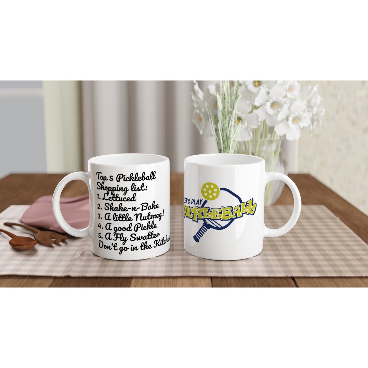 Two white ceramic 11oz mugs with original motto Top 5 Pickleball Shopping list lettuce, Shake-n-bake, Nutmeg, Pickle, a Fly Swatter Don’t go in the kitchen front side and Let's Play PickleBall logo on back coffee mugs are dishwasher and microwave safe from WhatYa Say Apparel sitting on coffee table with green and white placemat.