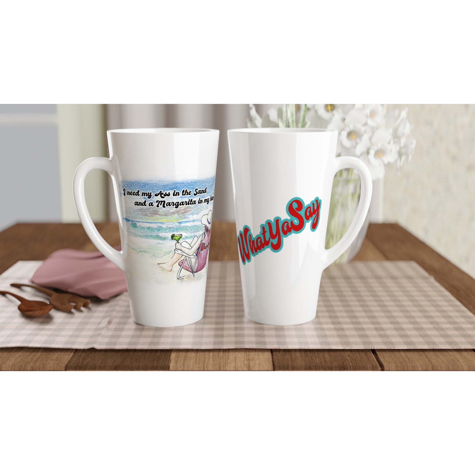 Two Seventeener white ceramic 17oz mugs with original motto I need my Ass in the Sand and a Margarita in my hand on front and WhatYa Say logo on back dishwasher and microwave safe from WhatYa Say Apparel sitting on coffee table with green and white placemat. 