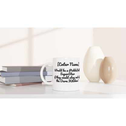 Personalized white ceramic 11oz mug with motto Personalized Your Name Would be a PickleBall Superstar if they could stay out of the Darn Kitchen! on front and Let's Play Pickleball logo on back dishwasher and microwave safe from WhatYa Say Apparel sitting on coffee table with books and two vases.