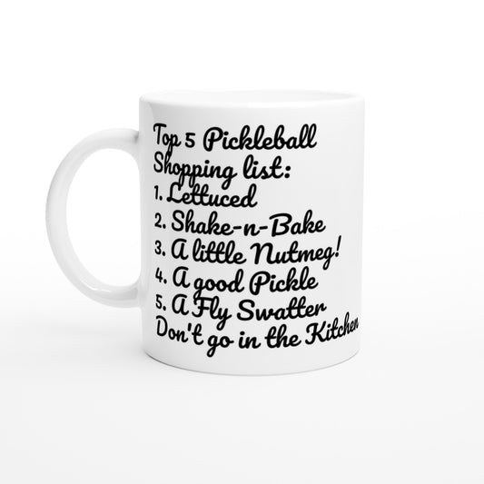 White ceramic 11oz mug with white handle original motto Top 5 Pickleball Shopping list lettuce, Shake-n-bake, Nutmeg, Pickle, a Fly Swatter Don’t go in the kitchen front side and Let's Play PickleBall logo on back dishwasher and microwave safe ceramic coffee mug from WhatYa Say Apparel front view.