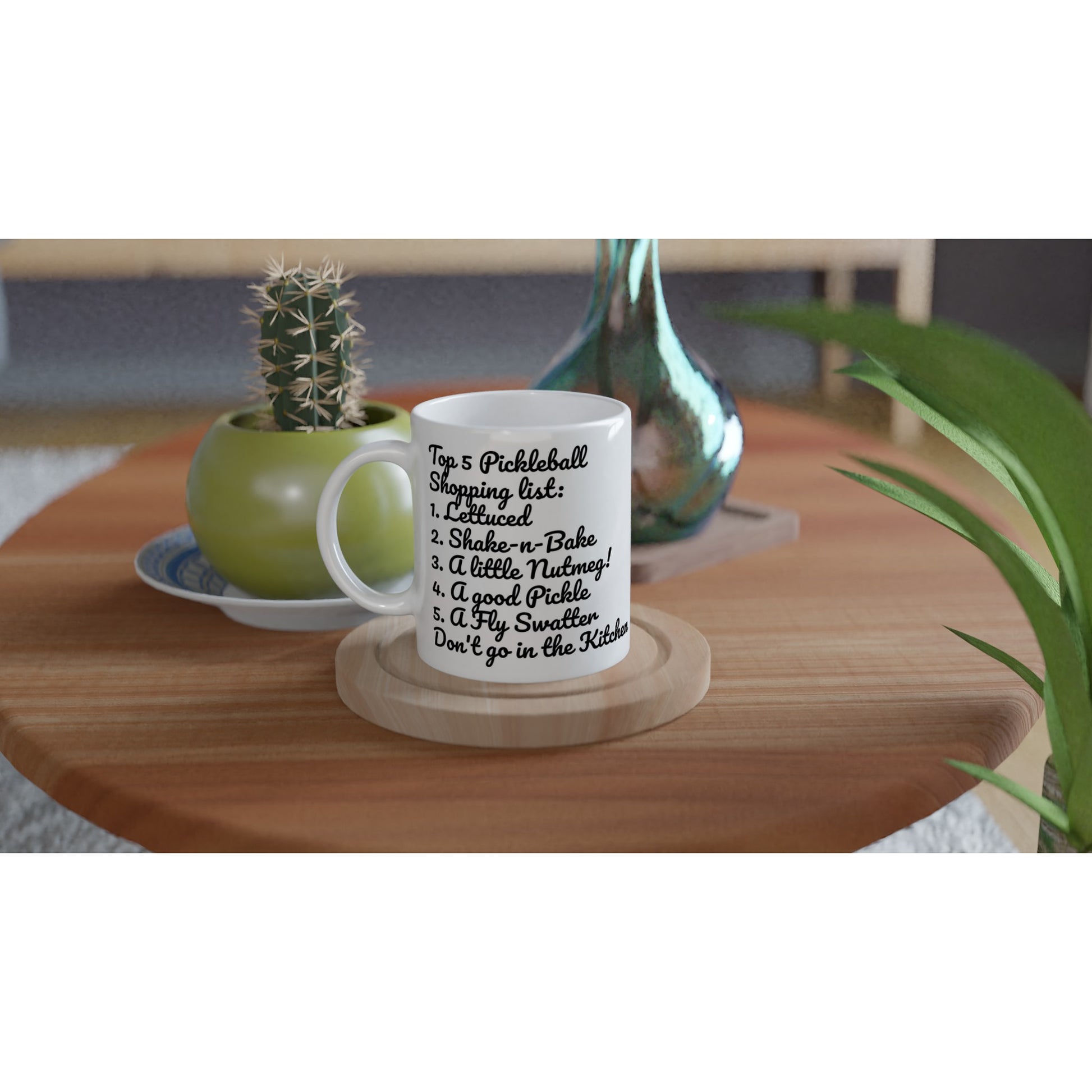 White ceramic 11oz mug with original motto Top 5 Pickleball Shopping list lettuce, Shake-n-bake, Nutmeg, Pickle, a Fly Swatter Don’t go in the kitchen front side and Let's Play Pickleball logo on back coffee mug dishwasher and microwave safe from WhatYa Say Apparel sitting on coaster on coffee table with green potted cactus and silver vase.